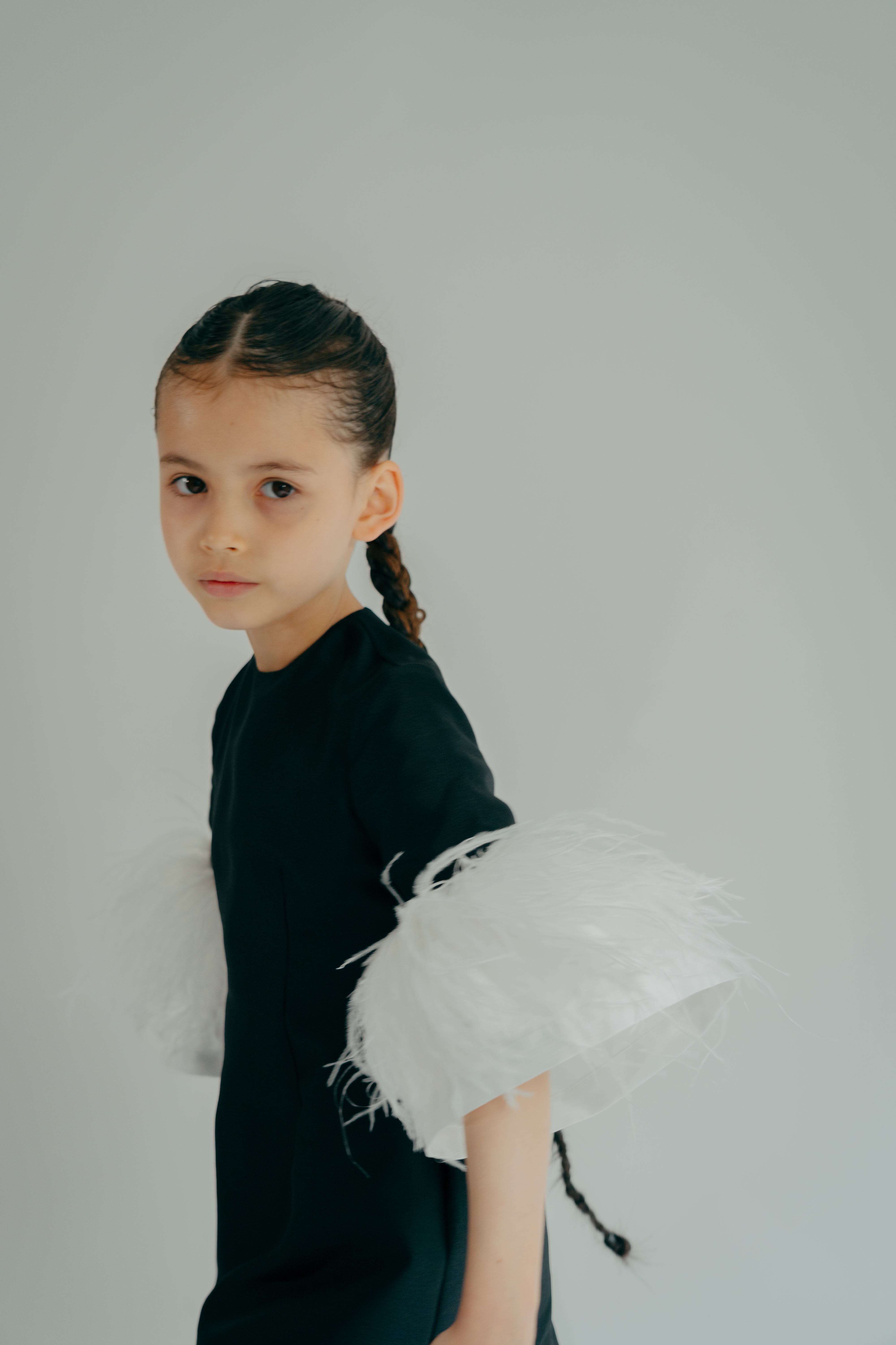 A young girl wearing a black A-line dress with white feathers attached to the sleeves.