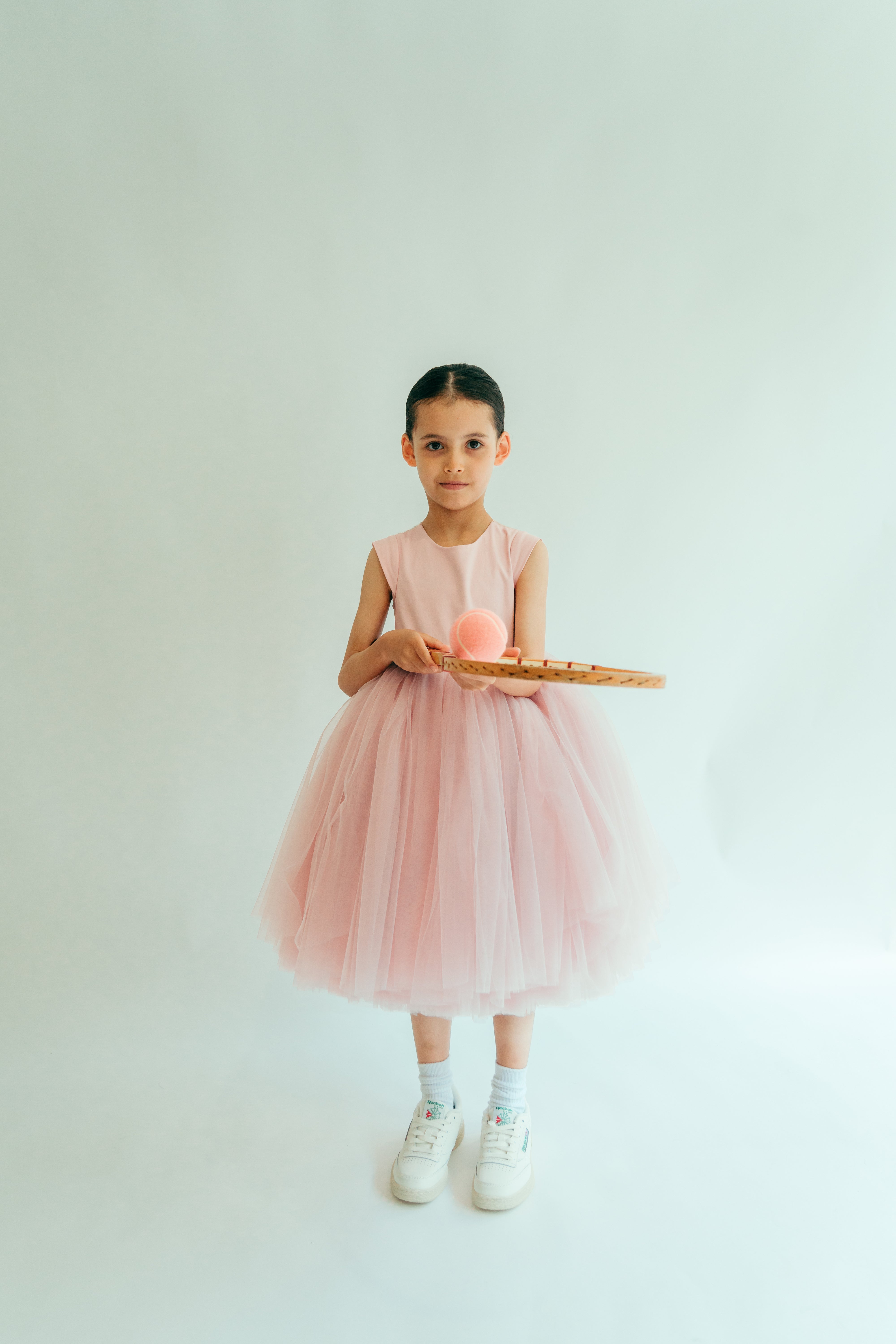 A young girl in a pink dress with sequins and pearl buttons, holding a tennis racket and ball.